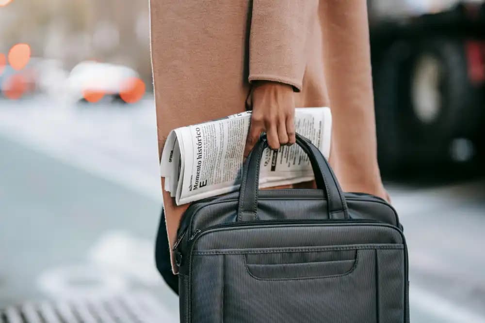 A person in a beige coat is holding a black bag and a folded newspaper while walking along a city street.